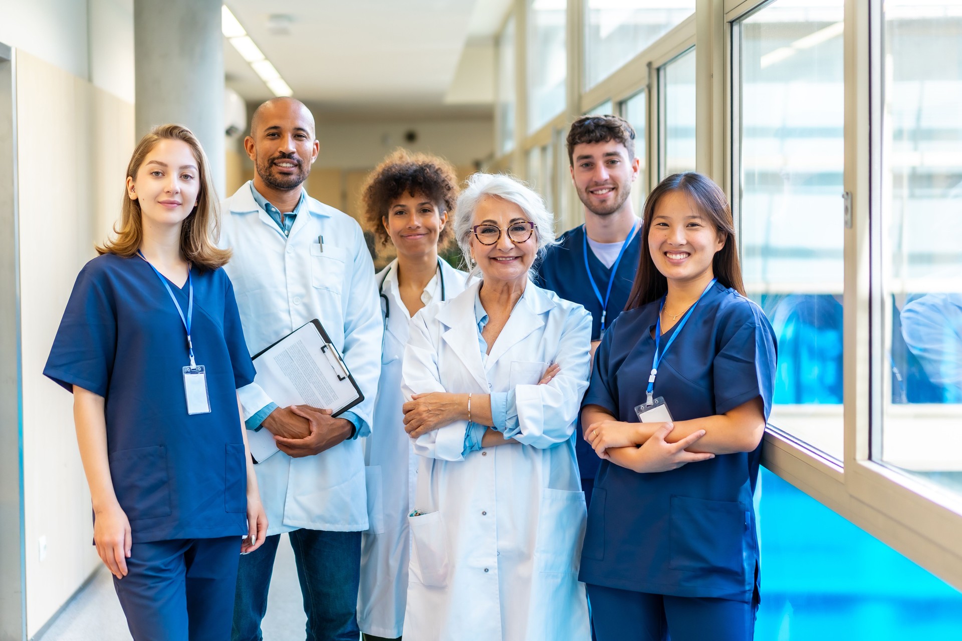 Group of diverse healthcare professionals in scrubs and lab coats, smiling and standing together in a hospital corridor, representing teamwork and care in the medical field.