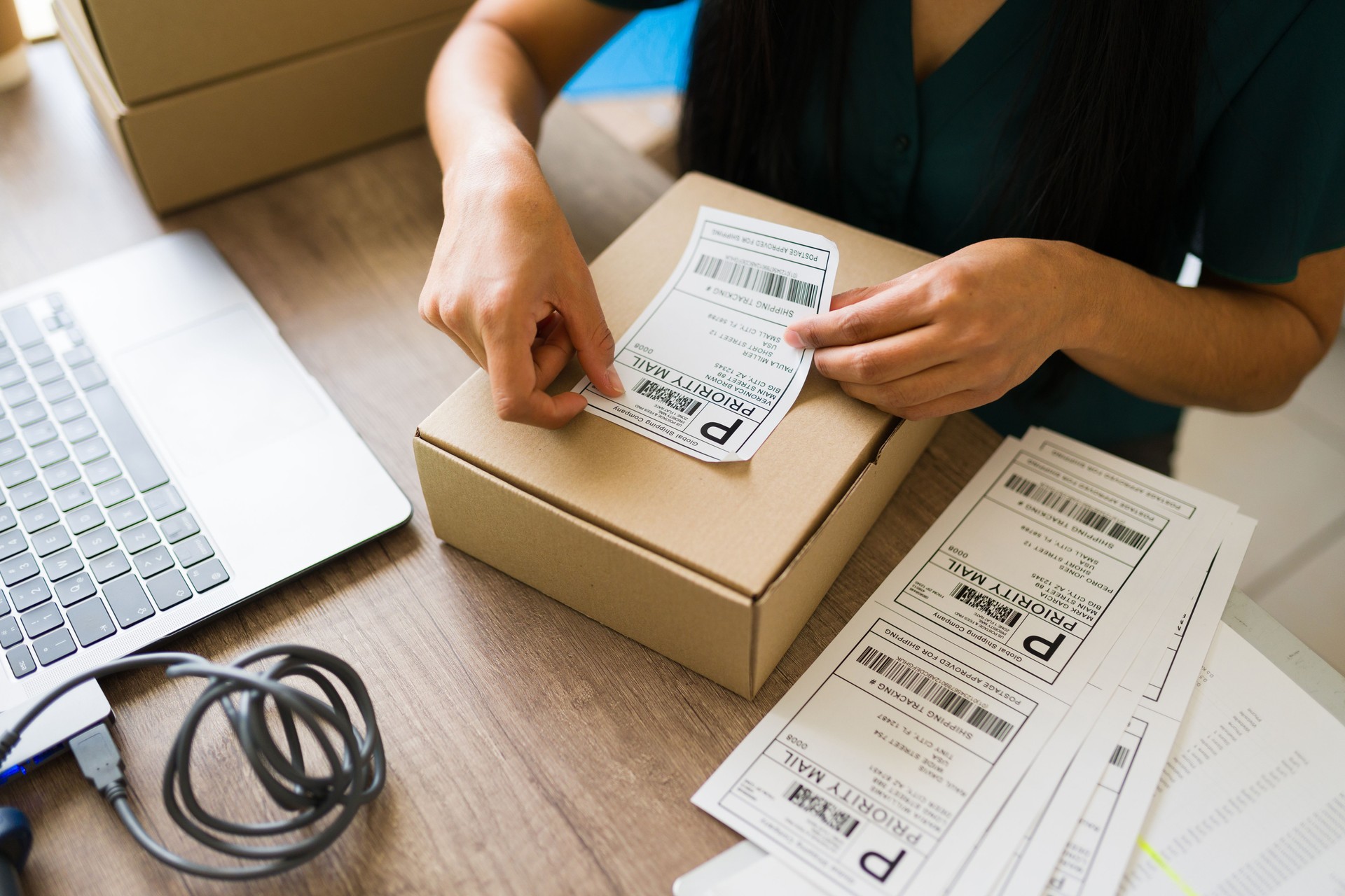 Person applying shipping label to a cardboard box on a desk with a laptop and multiple shipping labels, illustrating local business logistics and e-commerce fulfillment.