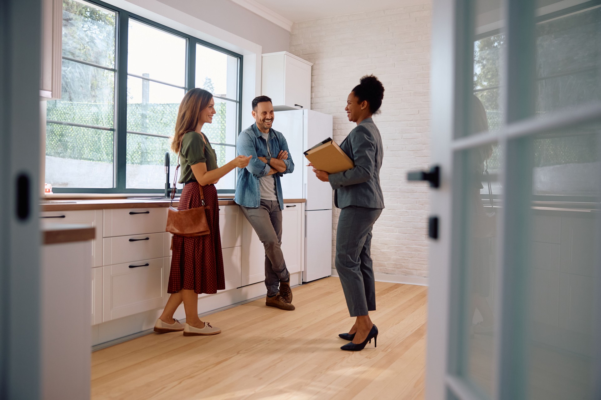 Three individuals engaged in conversation in a modern kitchen, featuring a woman in a green top and red skirt, a man in a denim jacket, and a professional woman in a suit holding a clipboard, emphasizing local business interactions.